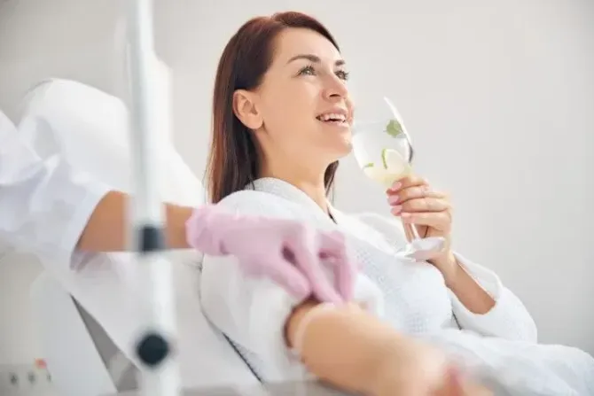 Woman in a white robe drinking water while receiving an IV infusion.