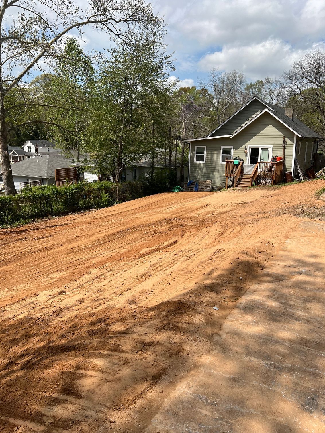 Dirt road leading to a small, weathered house. Trees and other houses in the background under a cloudy sky.