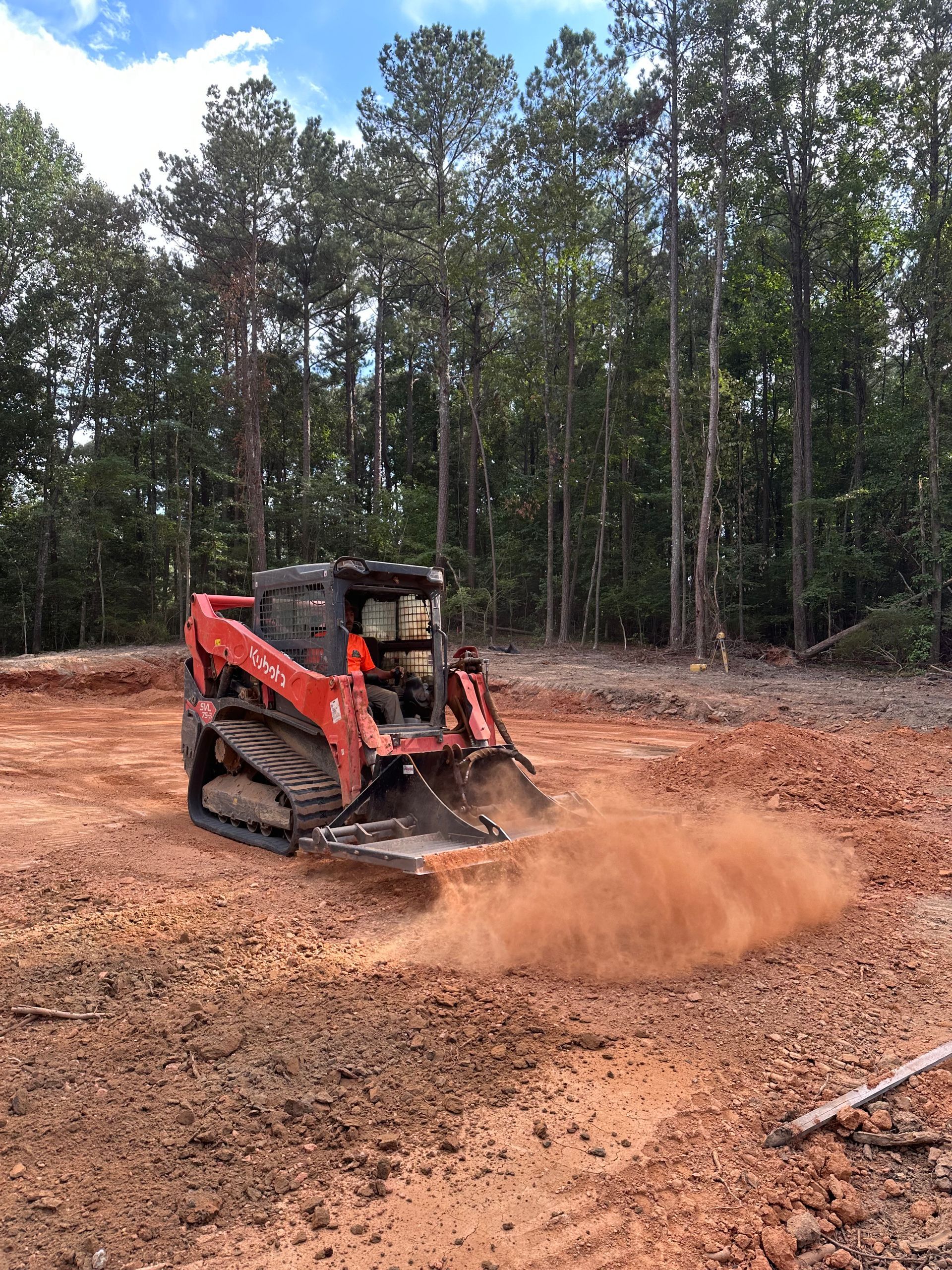 Red skid steer tractor kicking up dirt on a cleared construction site with trees in the background.