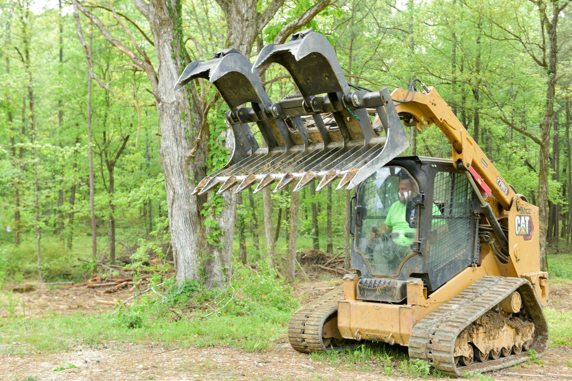 Yellow skid-steer loader with grapple attachment in wooded area. Operator visible inside cab.