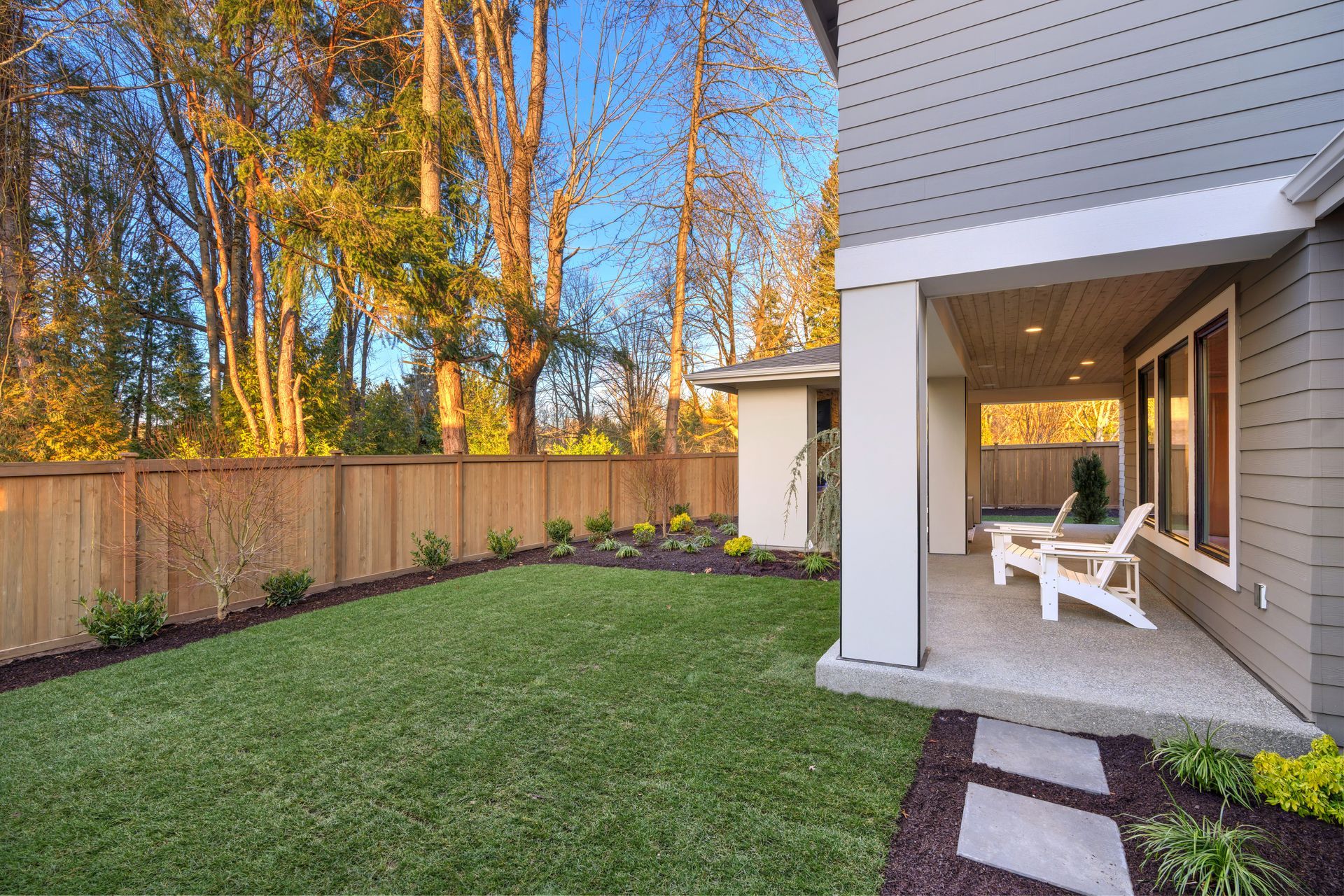 Backyard with green lawn, wooden fence, porch with chairs, and trees.