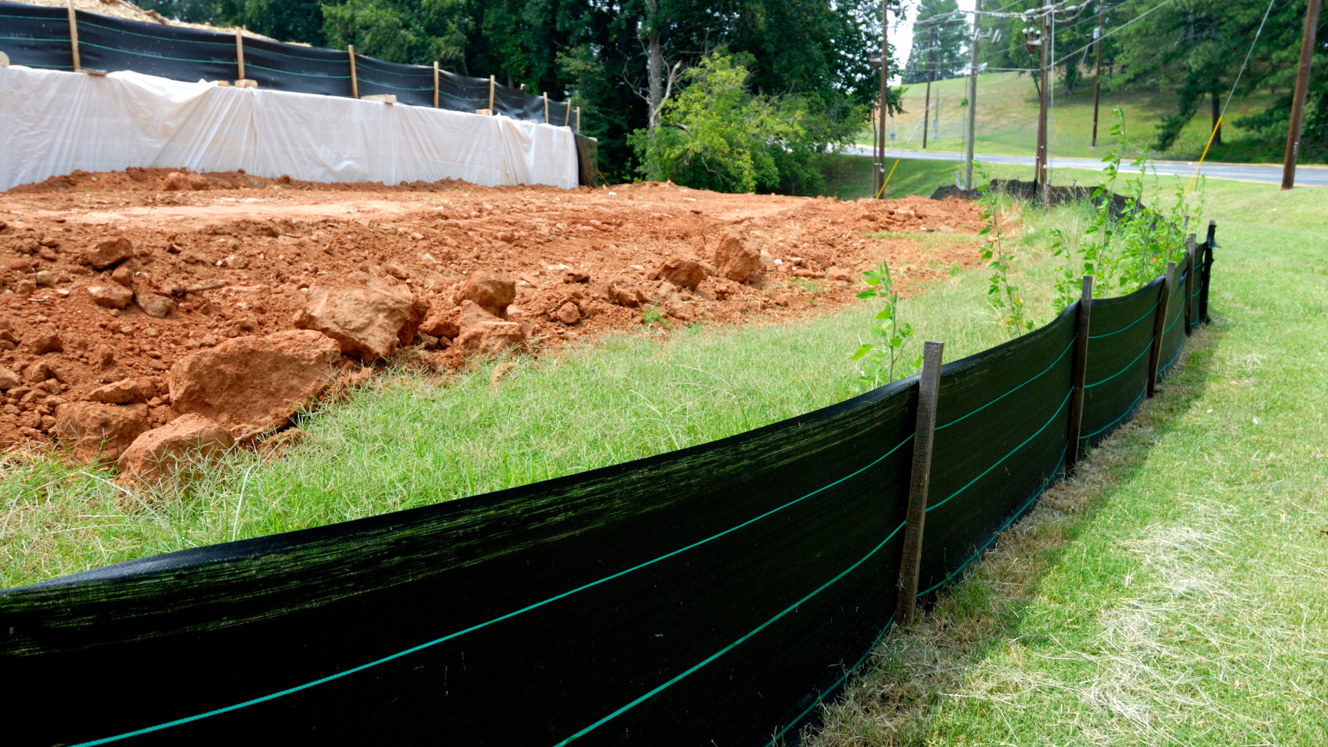 A construction site protected by a black silt fence on a grassy area, with exposed red soil.