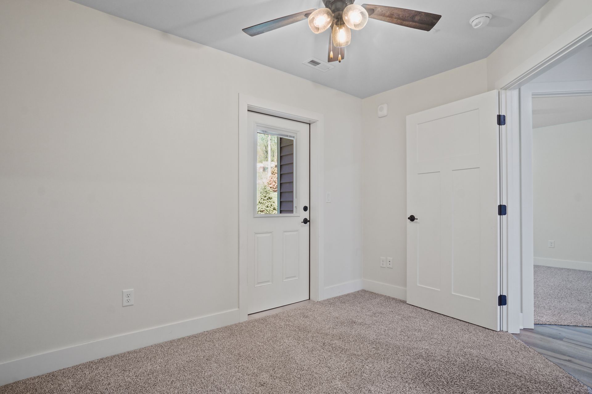 Empty white bedroom with carpeted floor, ceiling fan, and two doors, one with a narrow window.