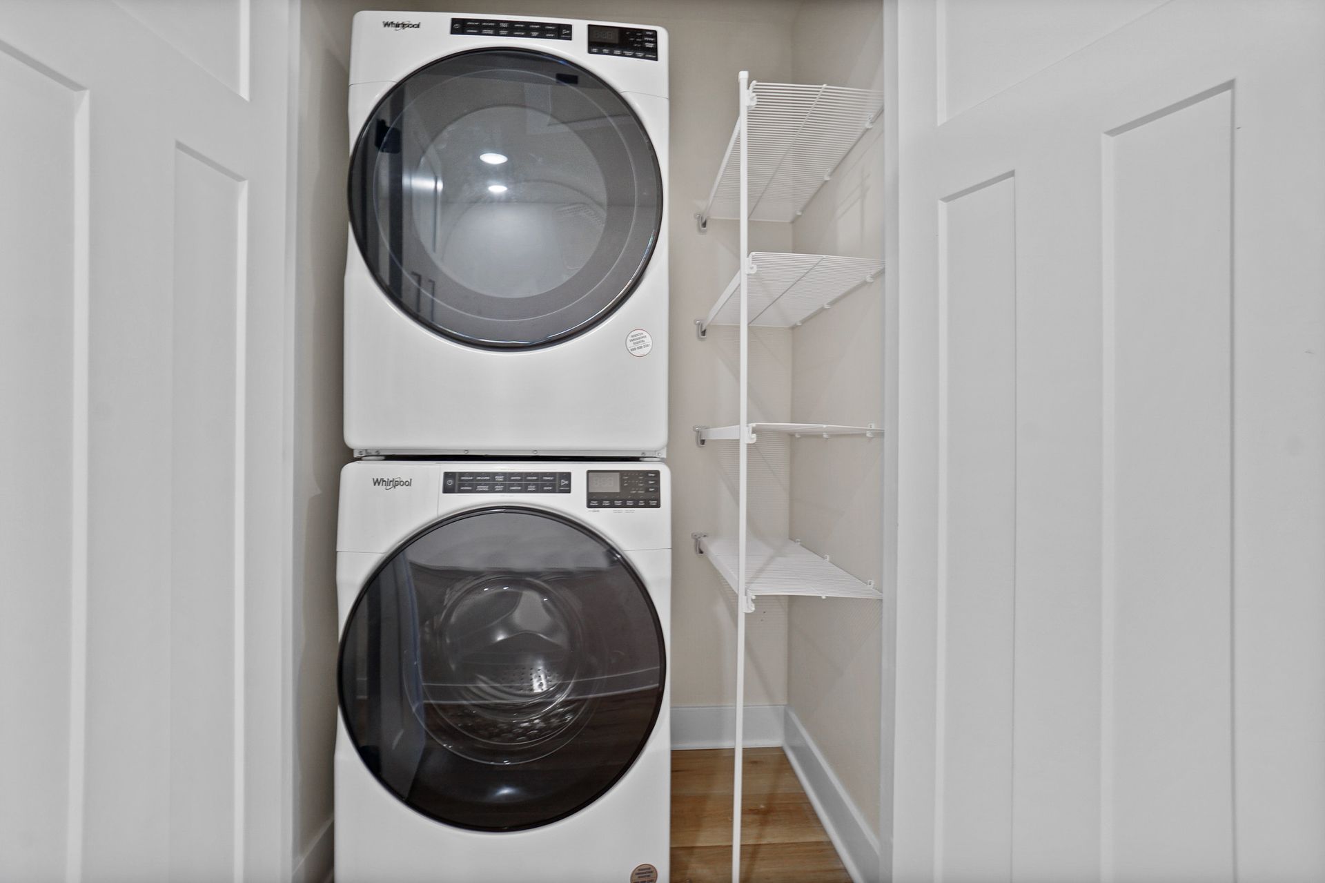 Stacked washer and dryer in a narrow laundry closet with white shelves and louvered doors