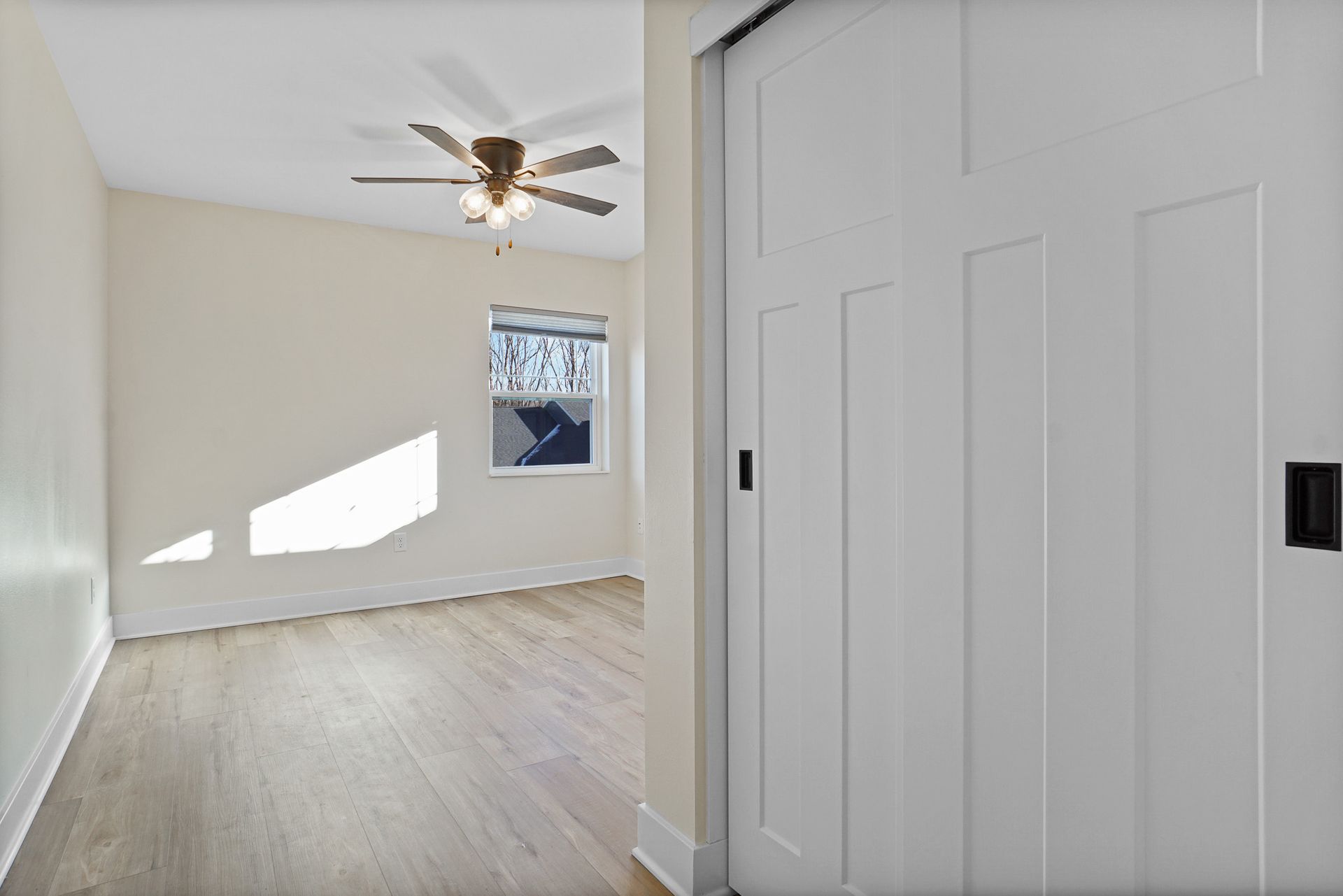 Empty bright room with light wood floor, ceiling fan, and a small window near a closet door