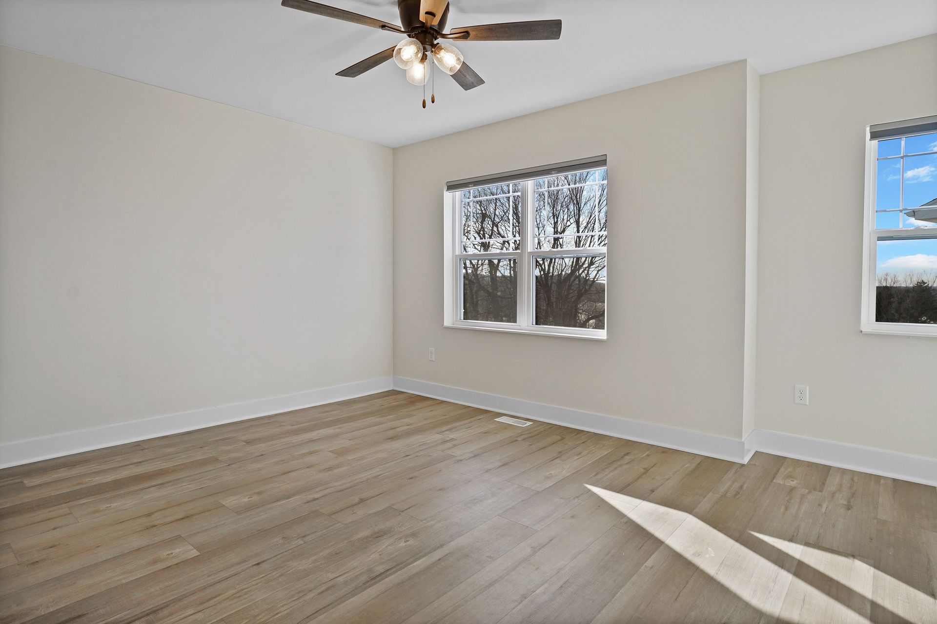 Empty beige bedroom with wood-look flooring, ceiling fan, and two windows letting in daylight