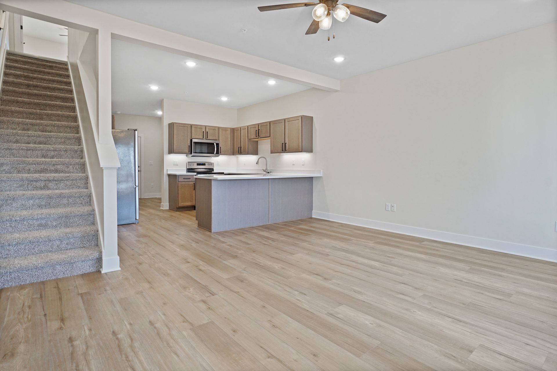 Empty living room with staircase, wood floors, and open kitchen with light cabinets