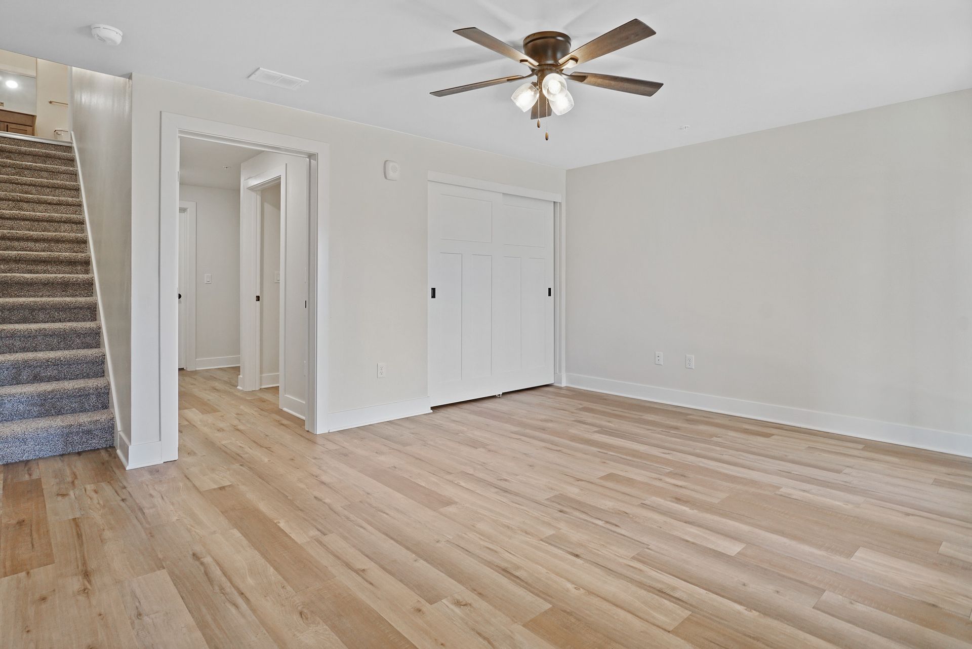 Empty living room with light wood floors, white walls, ceiling fan, and carpeted stairs at left