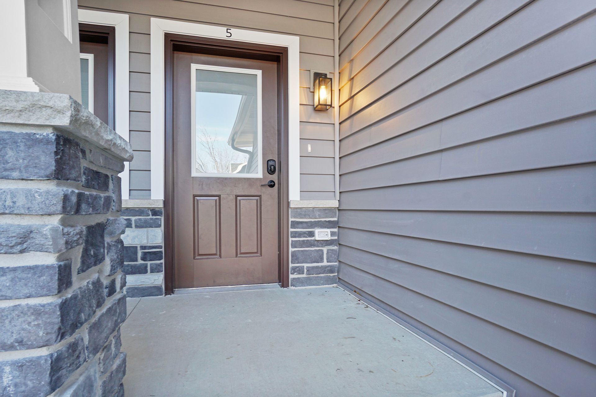 Front porch with tan front door, gray siding, stone columns, and a lit wall lantern