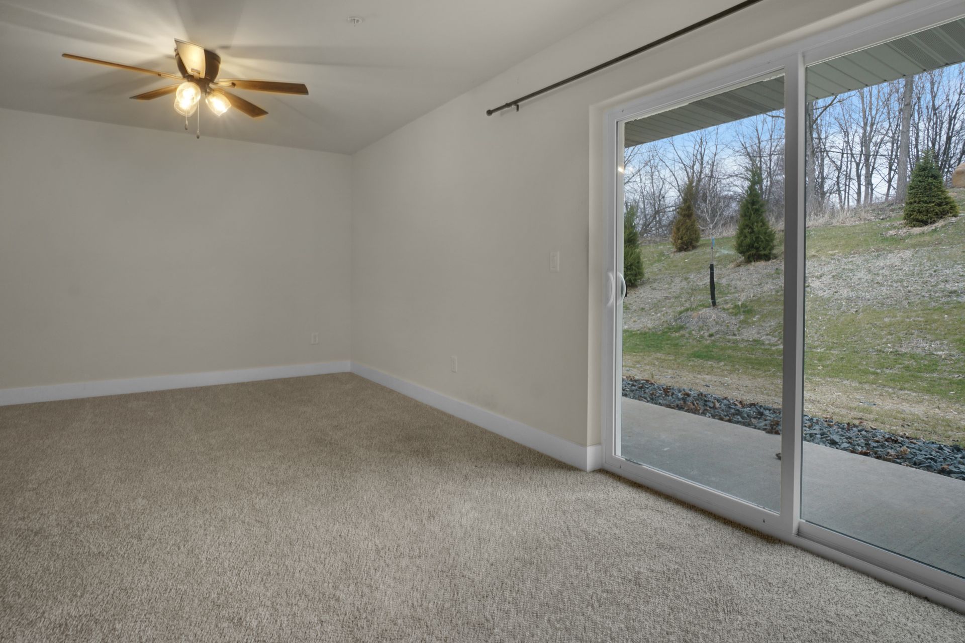 Empty beige room with ceiling fan, carpet, and sliding glass door to a grassy hillside outdoors