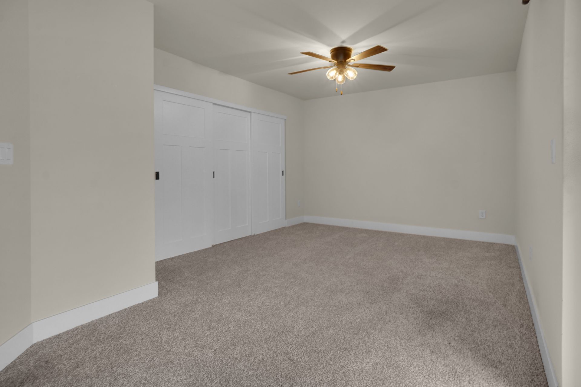 Empty beige bedroom with carpet, white closet doors, and a ceiling fan with light.