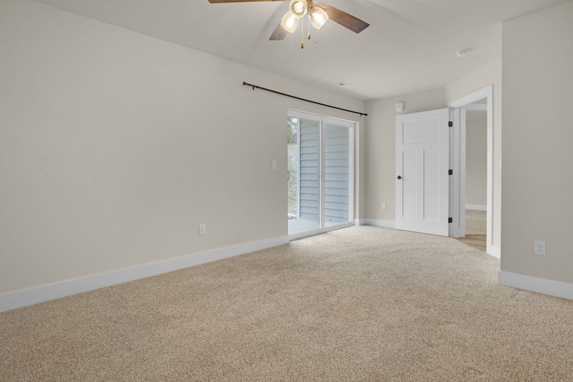Empty beige-carpeted bedroom with white walls, ceiling fan, and sliding glass doors to a balcony.