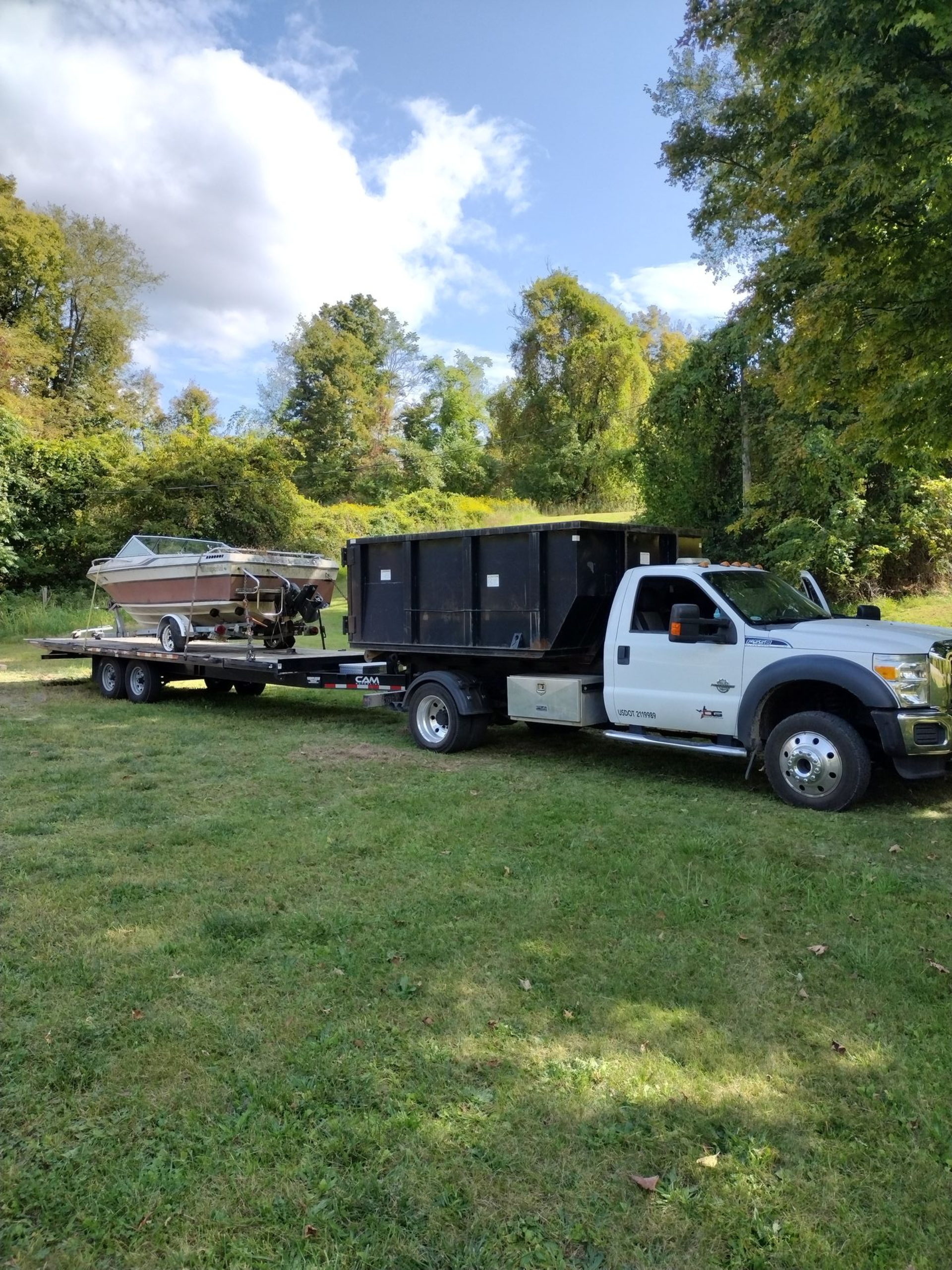 White truck with a dump bed towing a trailer with a boat in a grassy area.