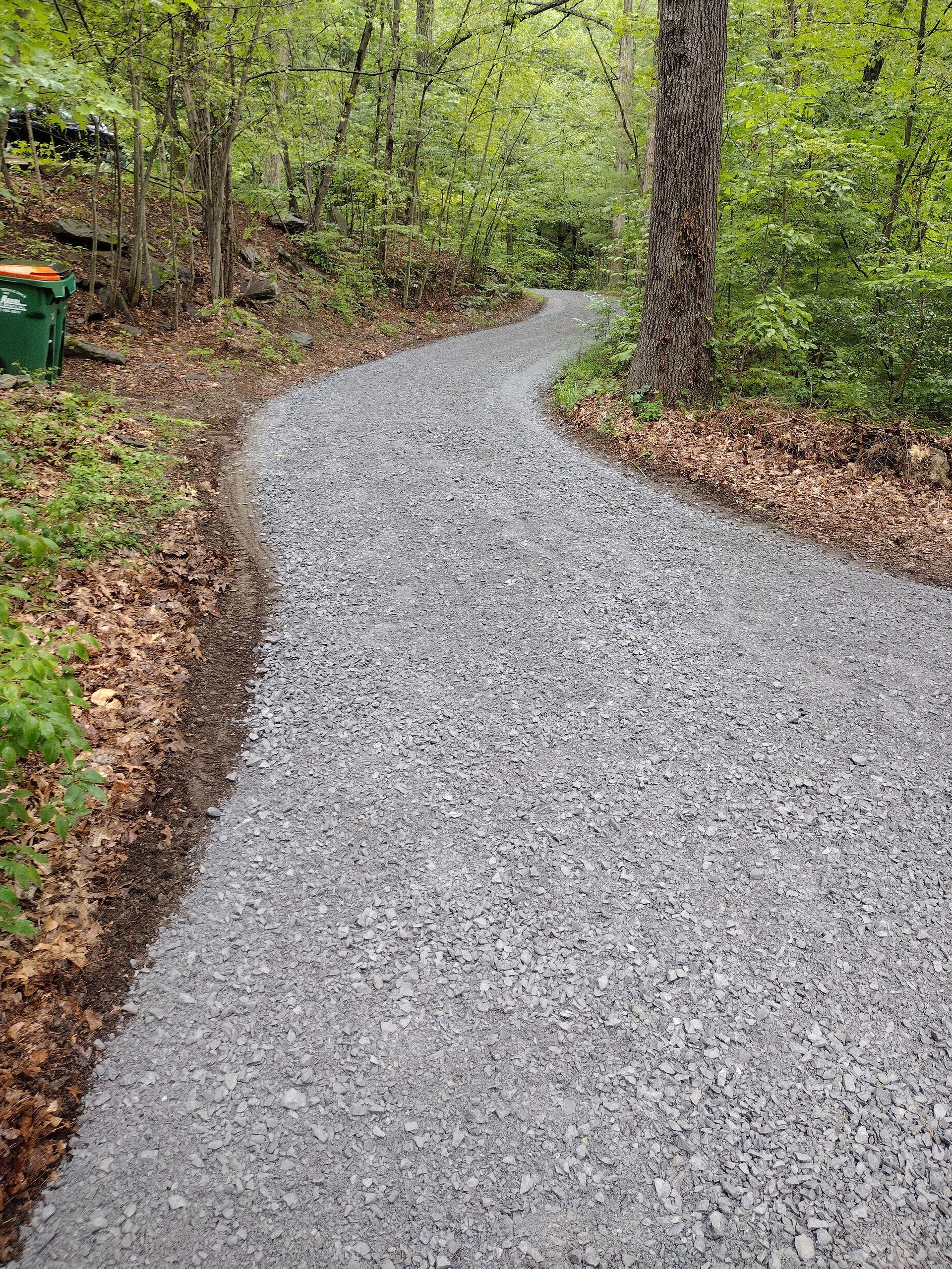 Gravel path curves through a wooded area. Edges of the trail are lined with leaves.