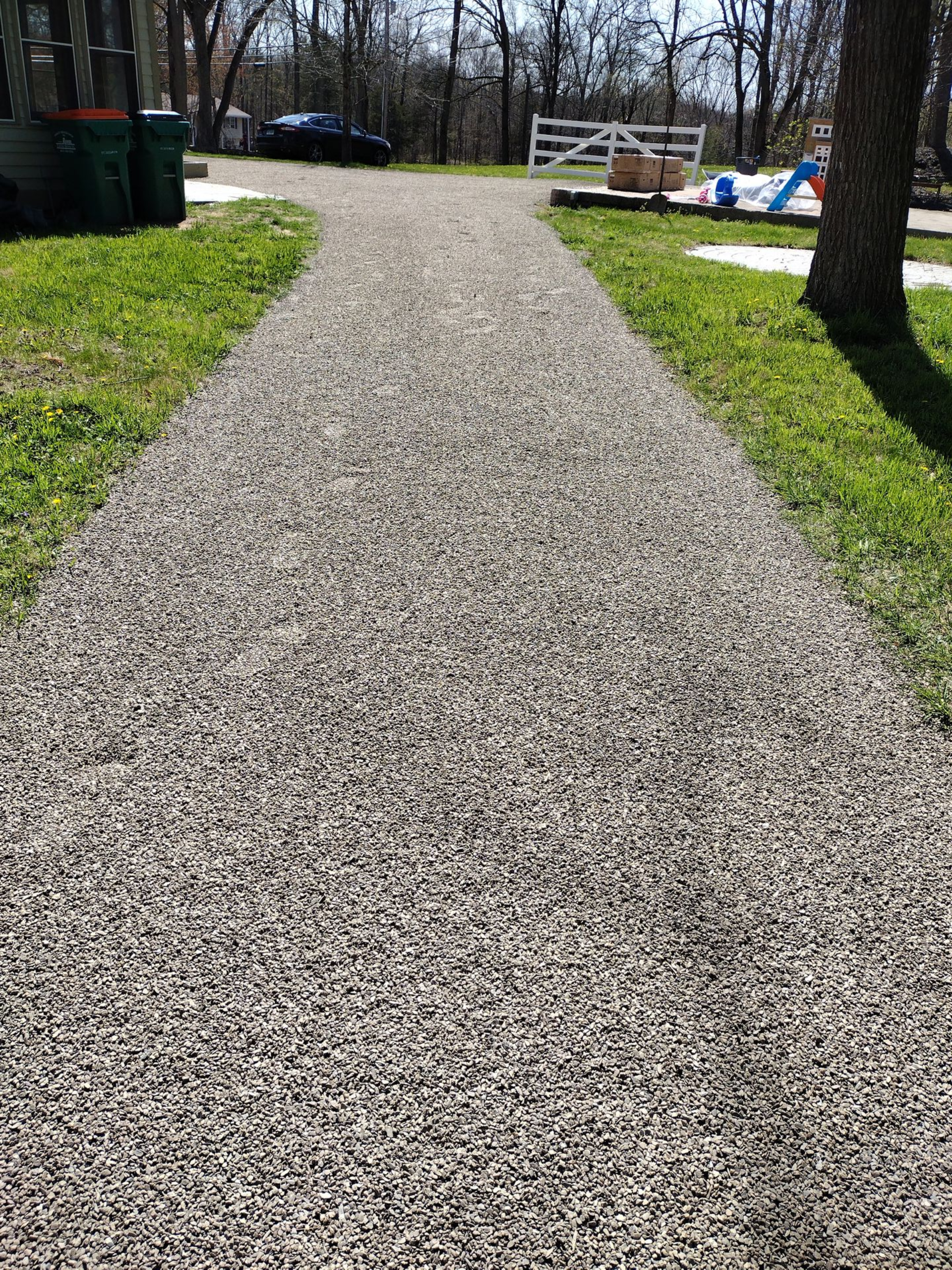 Gravel pathway through green grass, leading toward trees and a white fence on a sunny day.