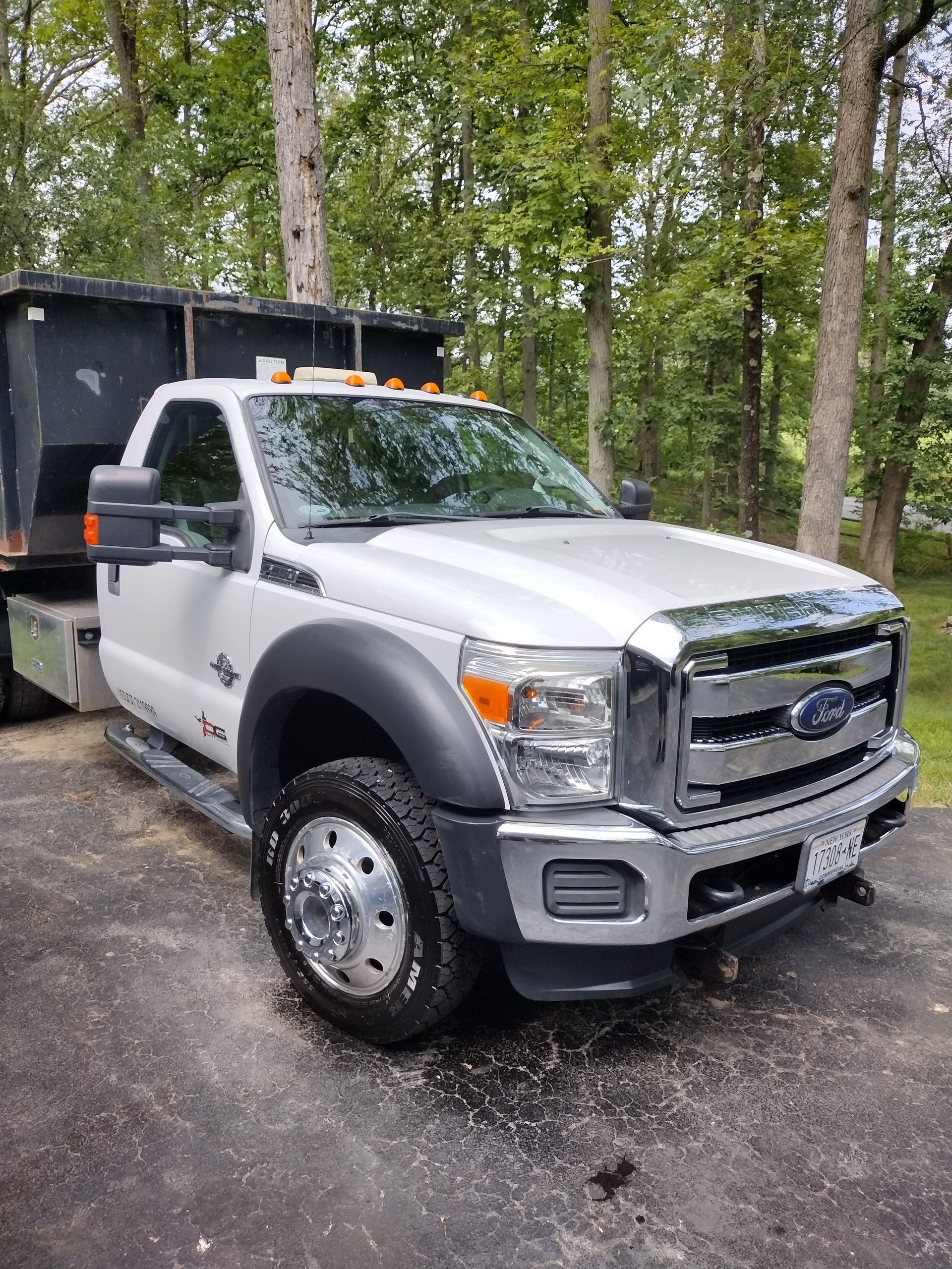 White Ford F-550 dump truck parked outside; black bed, chrome accents, and trees in background.
