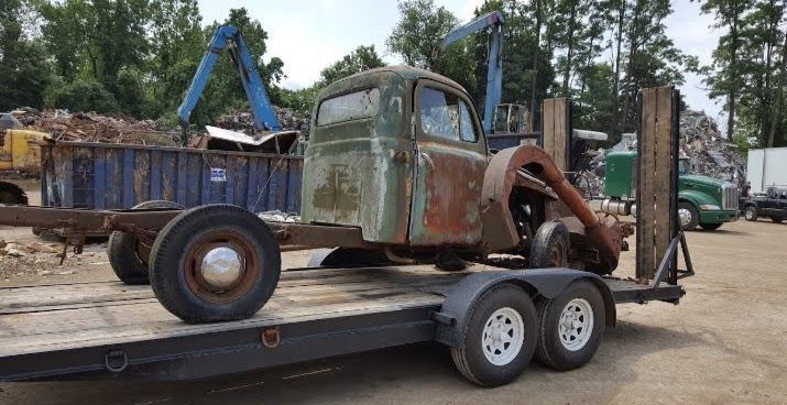Rusty truck cab on a trailer in a junkyard, blue container in background.