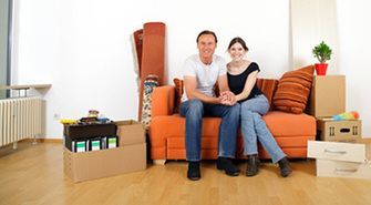 A man and a woman are sitting on a couch in a living room surrounded by cardboard boxes.