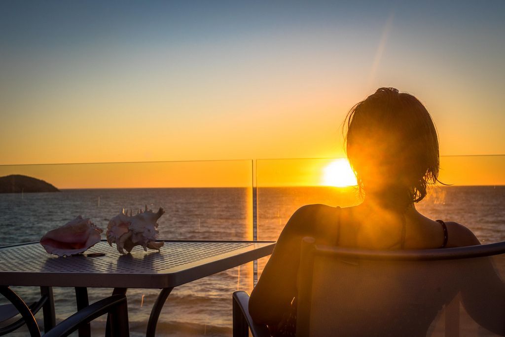A woman is sitting at a table looking at the sunset over the ocean.