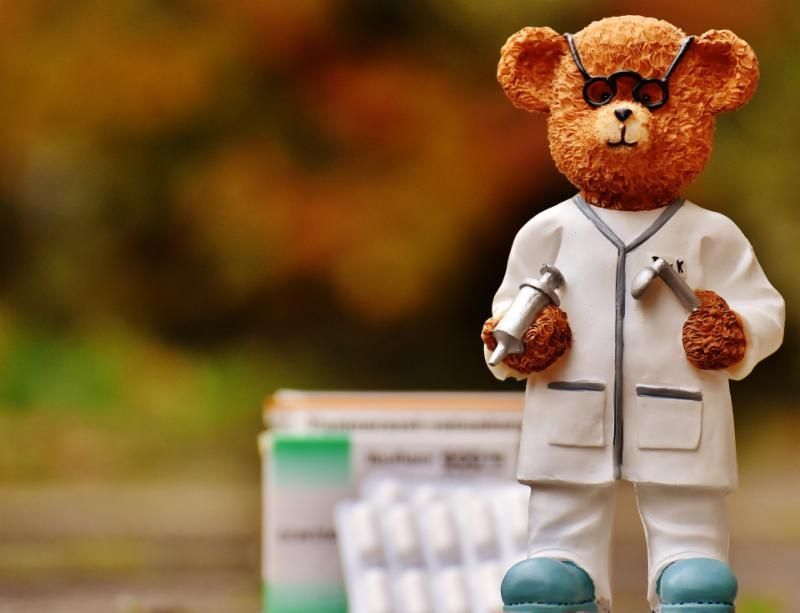 A teddy bear dressed as a doctor is holding a syringe and a stethoscope.