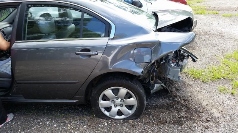 A gray car with a damaged side is parked in a gravel lot.