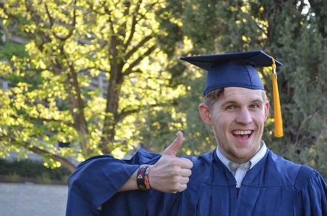 A man in a graduation cap and gown is giving a thumbs up.