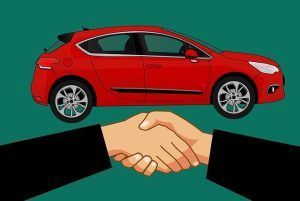 Two people are shaking hands in front of a red car.