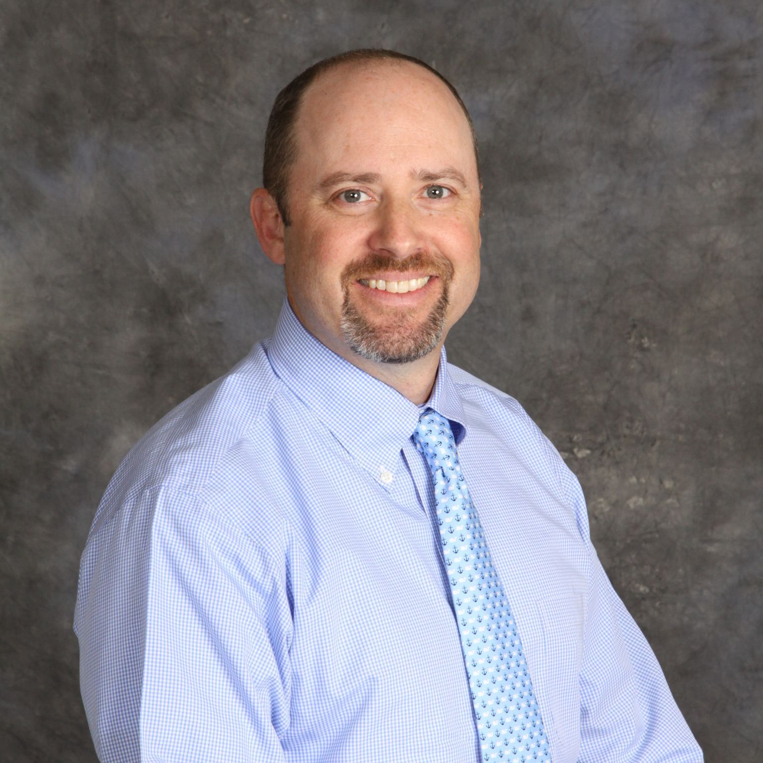 Man in blue striped shirt and tie smiles, posed against a gray backdrop.