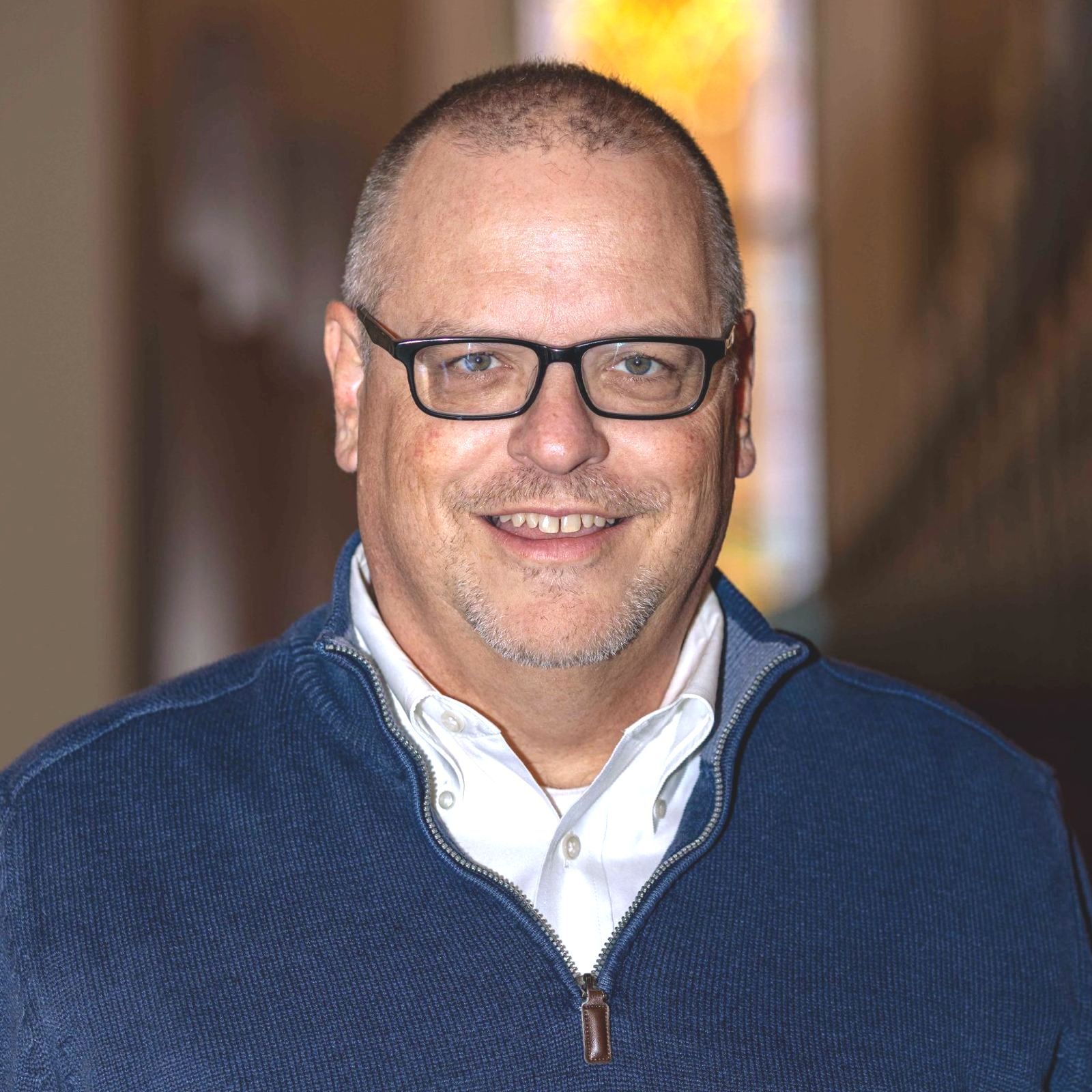 Man with glasses smiling, wearing a blue sweater over a white collared shirt, indoors.