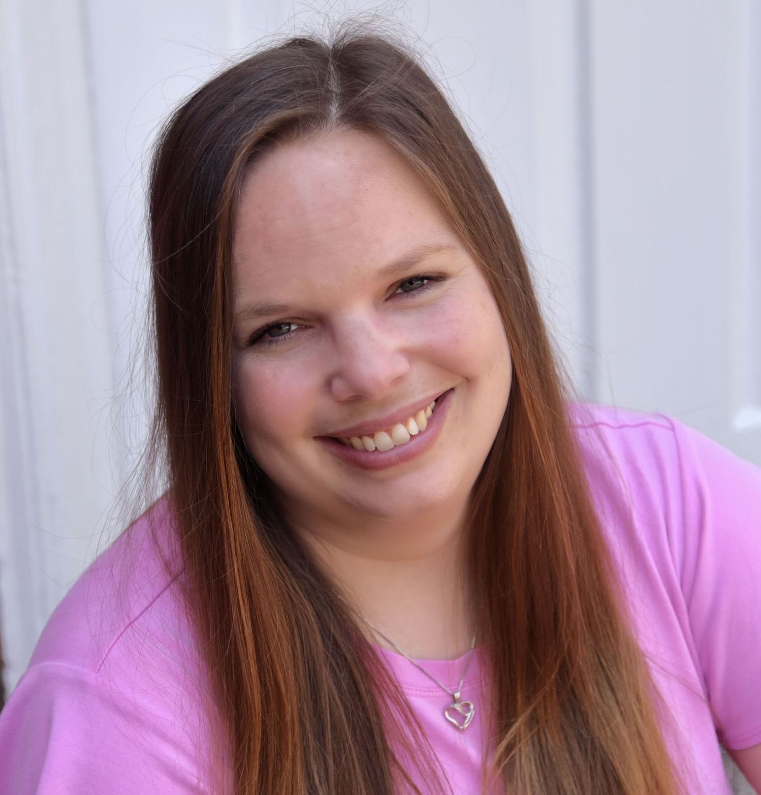 Woman with long brown hair smiles, wearing a pink shirt, in front of a white background.