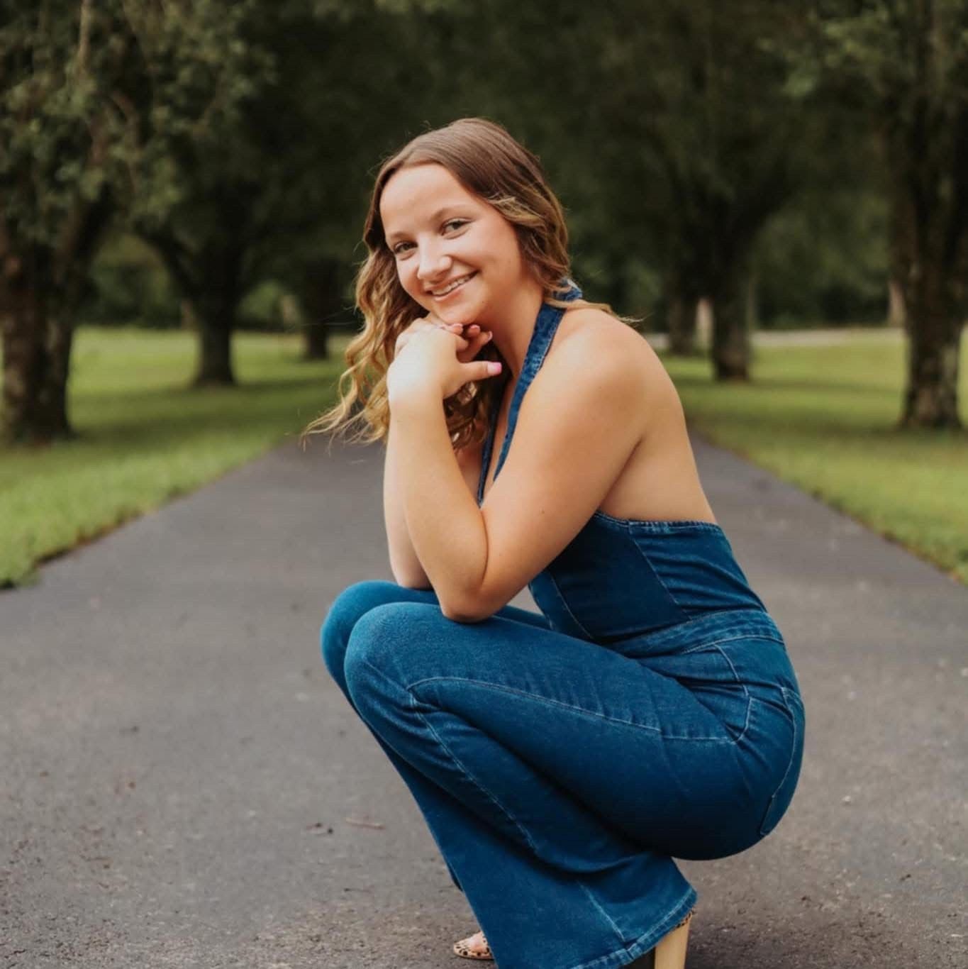 Woman in blue denim jumpsuit squats on a paved path, smiling, trees in background.