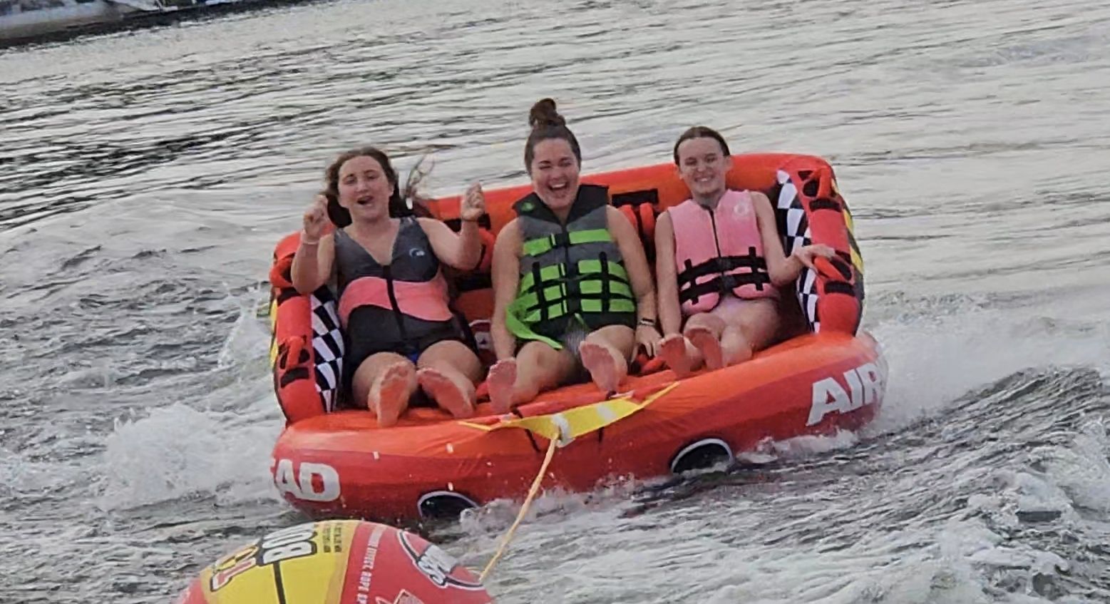 Three people in life vests on an inflatable tube being pulled by a boat on water; they are smiling and enjoying the ride.