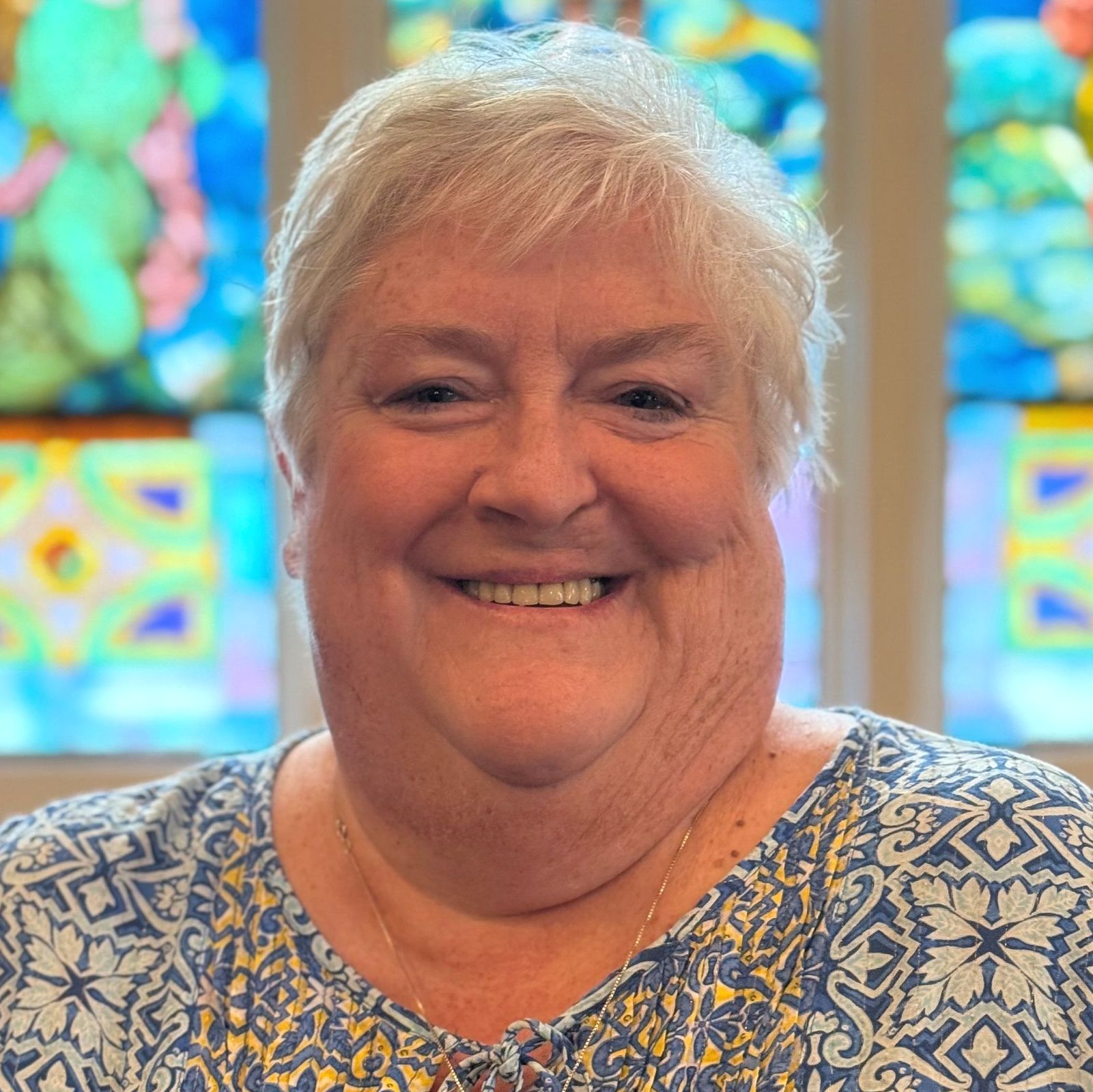Woman with short white hair smiles, wearing a blue patterned top, in front of stained glass.