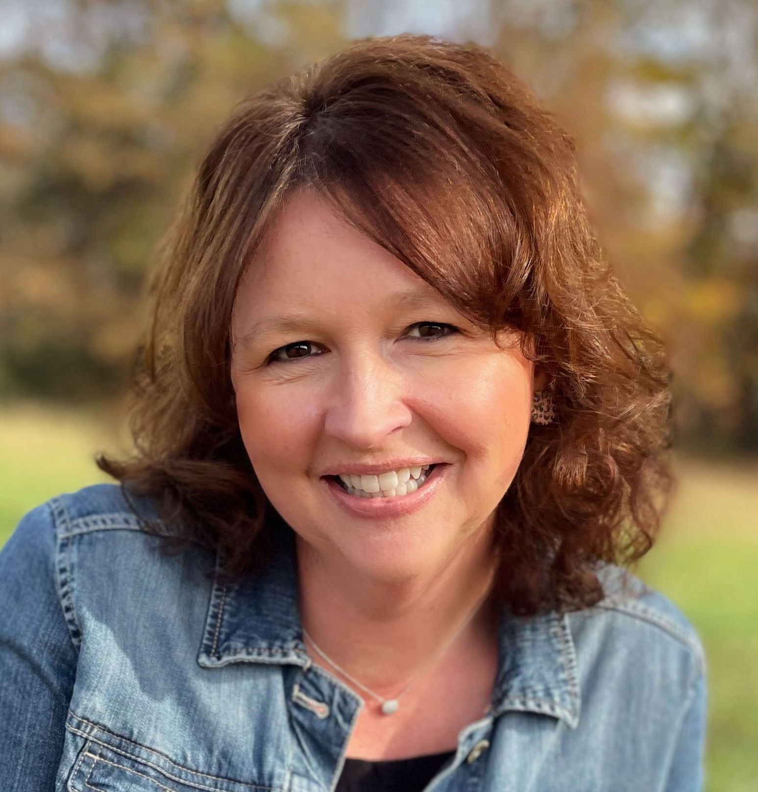 Woman smiling outdoors, wearing a denim jacket. Brown hair, blurred background of trees and grass.