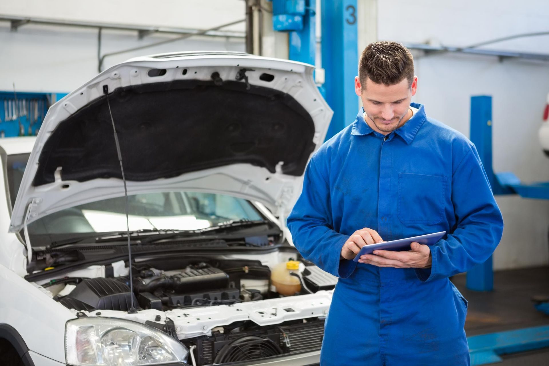Mechanic in blue coveralls using a tablet beside a car with its hood open in a repair shop