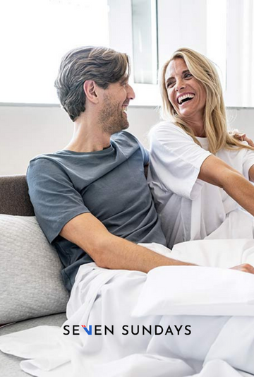 Zwei lächelnde Menschen sitzen auf einem Sofa und schauen sich in einem hellen Wohnzimmer an.