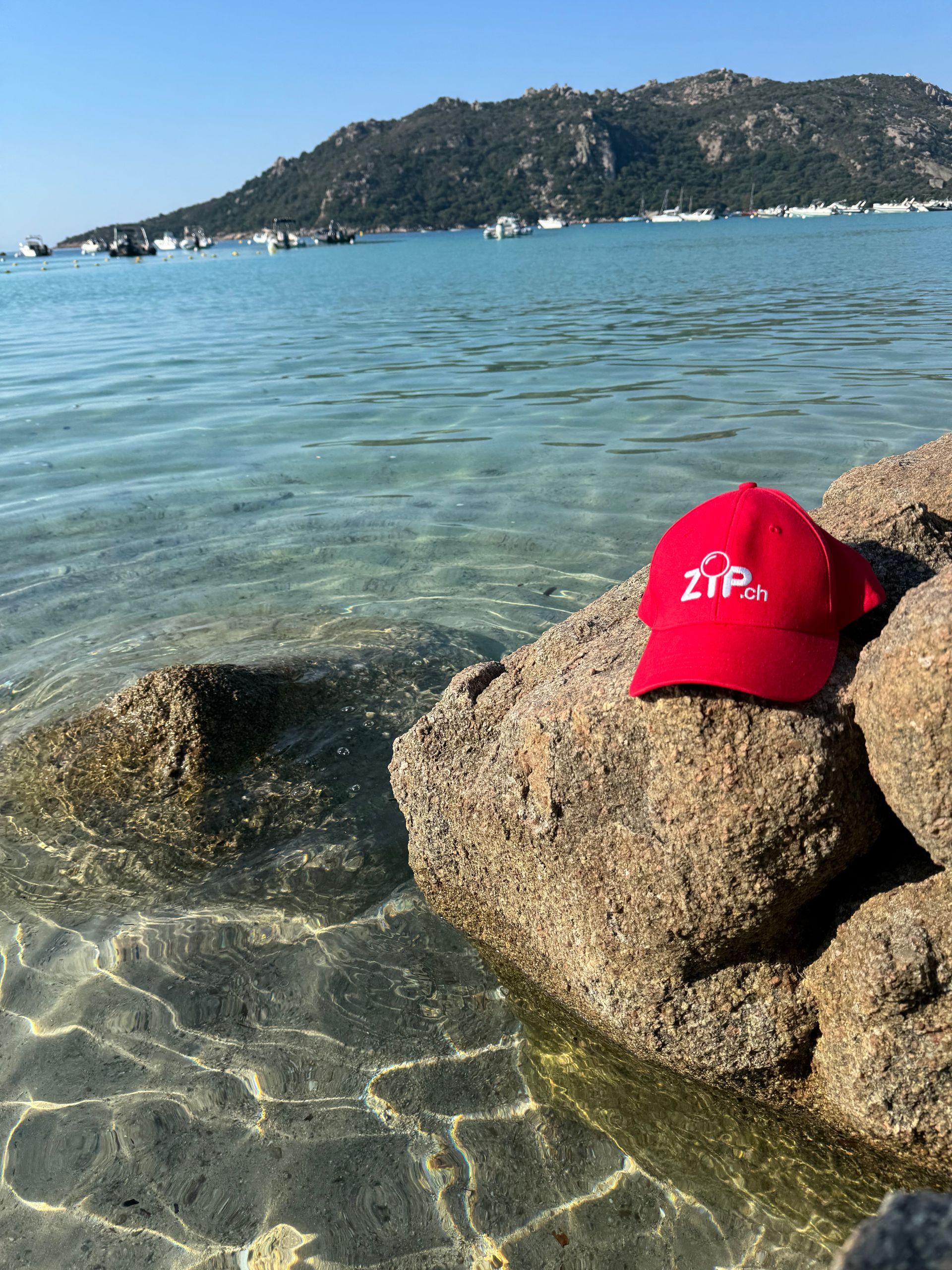 Red baseball cap on a rock by clear shallow water, with a distant tree-covered island in the background