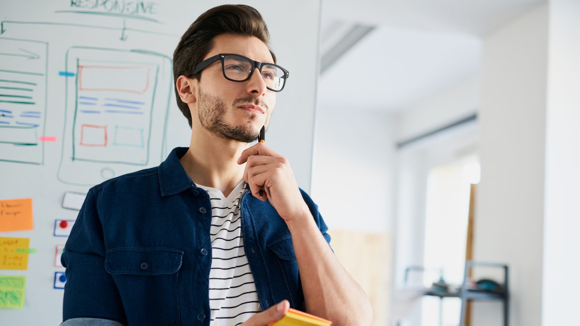 Man with glasses, looks up thoughtfully, holding pen near whiteboard with sketches in office.