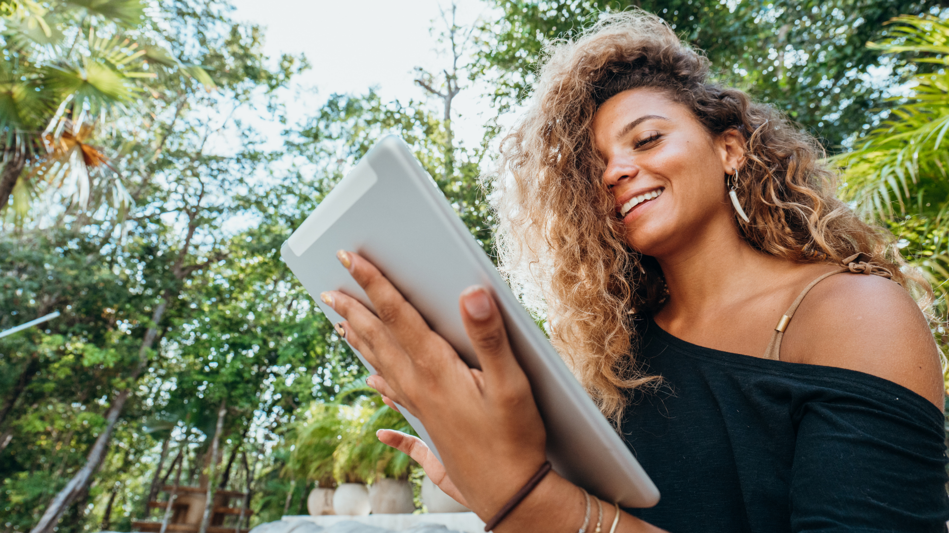 Woman with curly hair smiles while using a tablet outdoors, surrounded by green foliage.
