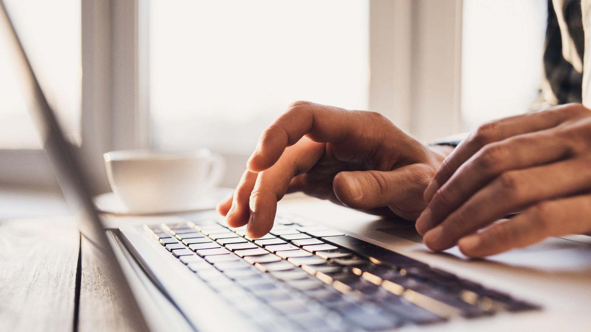 Hands typing on a laptop keyboard near a coffee cup by a bright window.