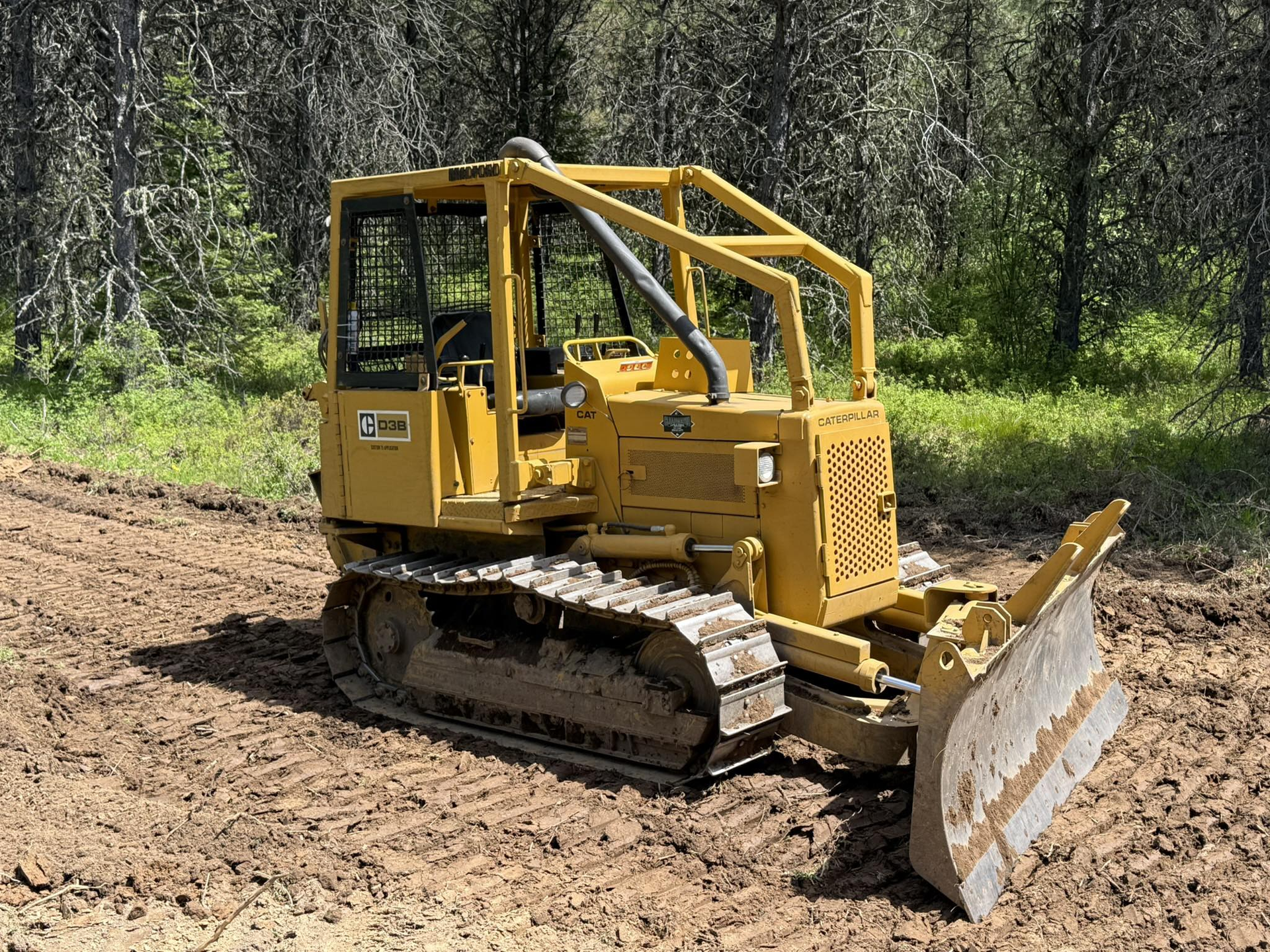 Yellow bulldozer with tracks and blade in a wooded area, sitting on a muddy path.