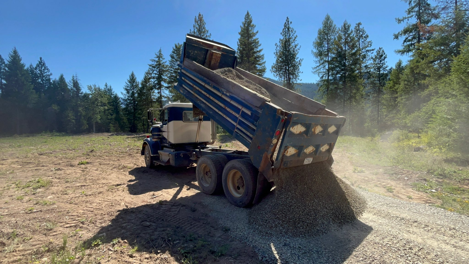 Dump truck unloading gravel on a dirt road in a sunny forest setting.