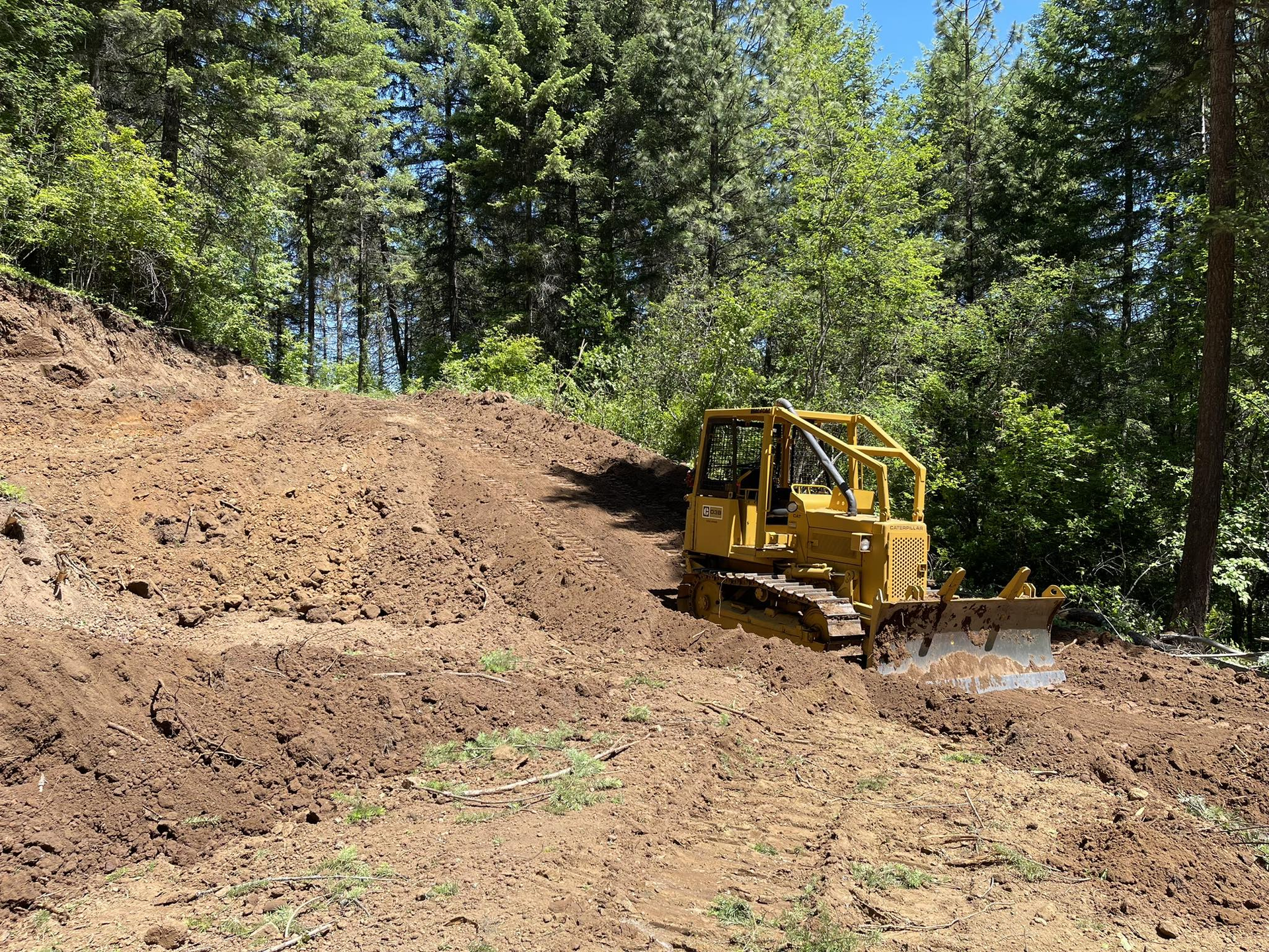 Yellow bulldozer on a muddy hillside, clearing land near trees under a bright sky.