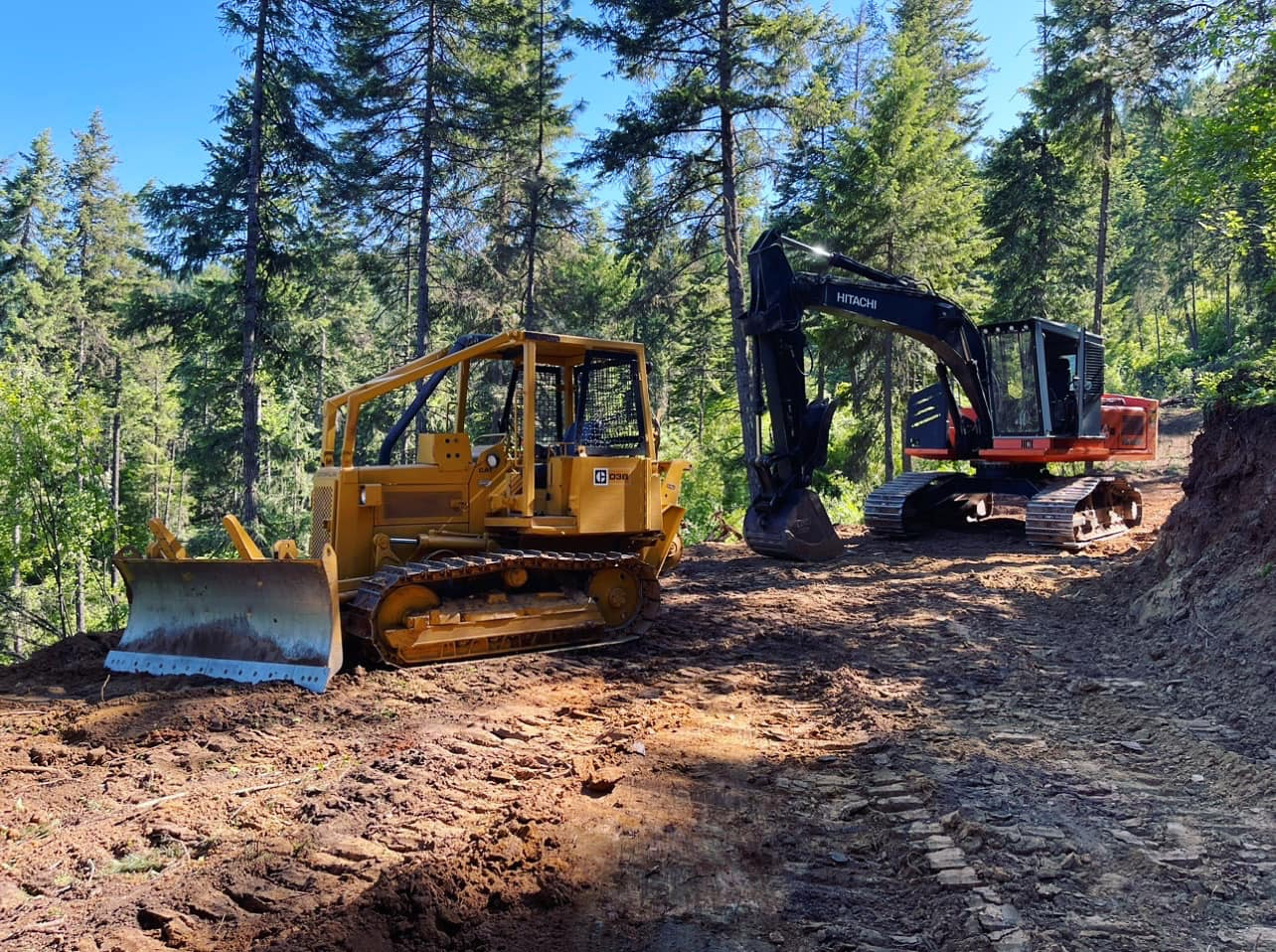 Yellow bulldozer and orange excavator on a dirt road in a forest.