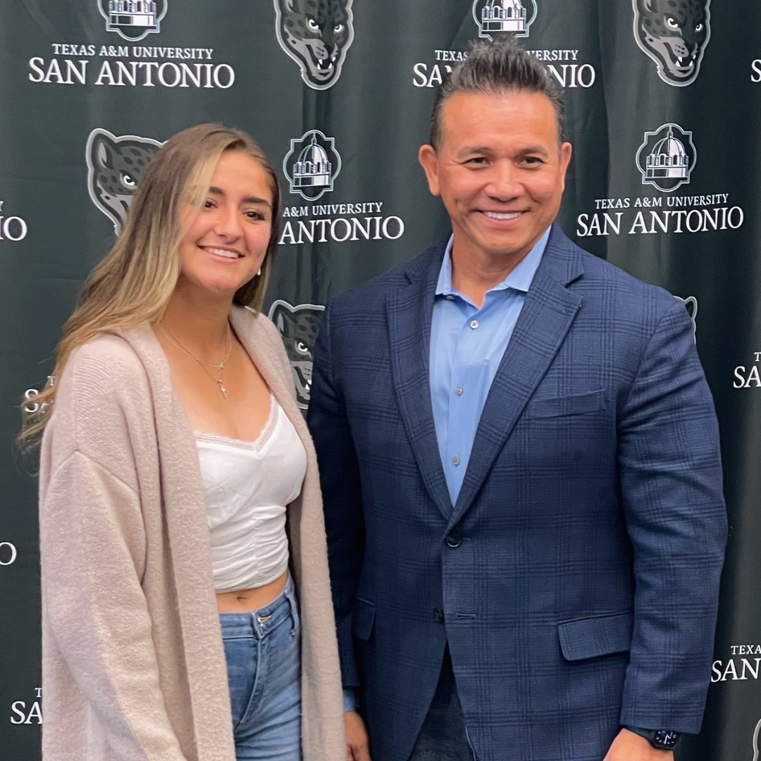 A person in a light cardigan and a person in a navy blazer stand smiling in front of a Texas A&M-San Antonio backdrop.