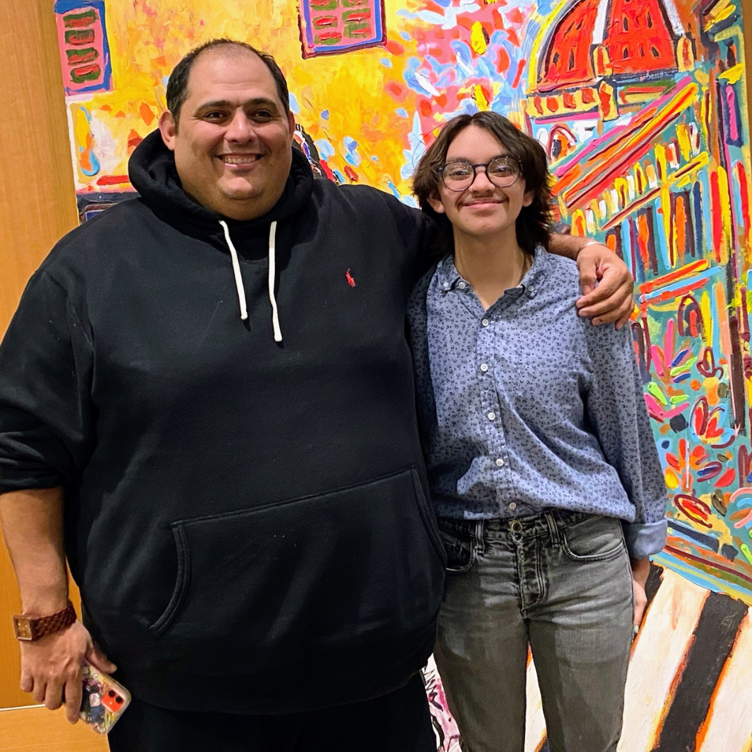 Two people pose smiling in front of a colorful, abstract painting of a city building with a domed roof.