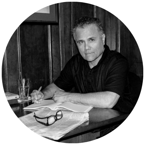 Black-and-white circular photo of a person writing in a notebook at a desk with a glass of water and glasses nearby.