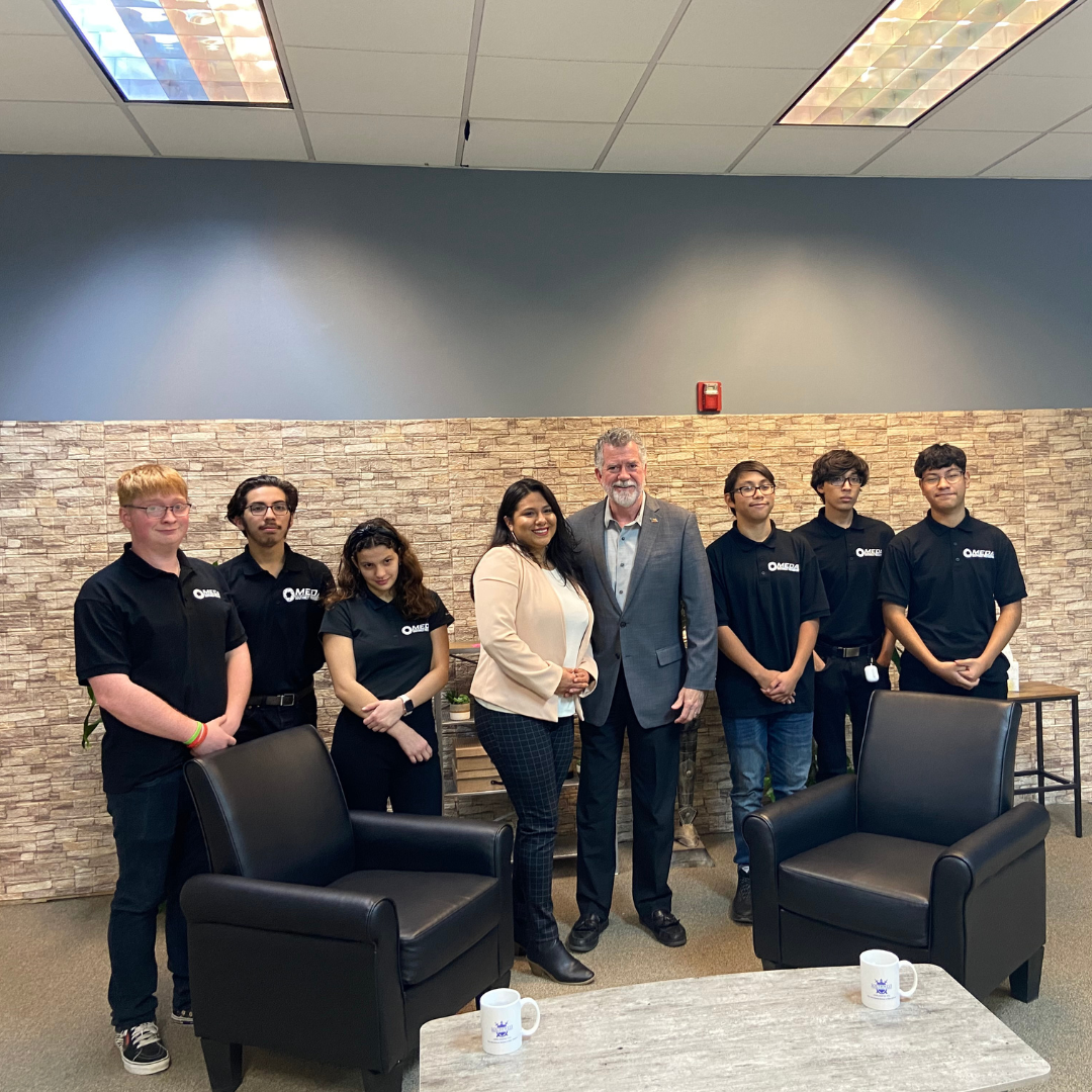A group of eight people stands in an office with a stone wall backdrop, two leather chairs, and a table with two mugs.