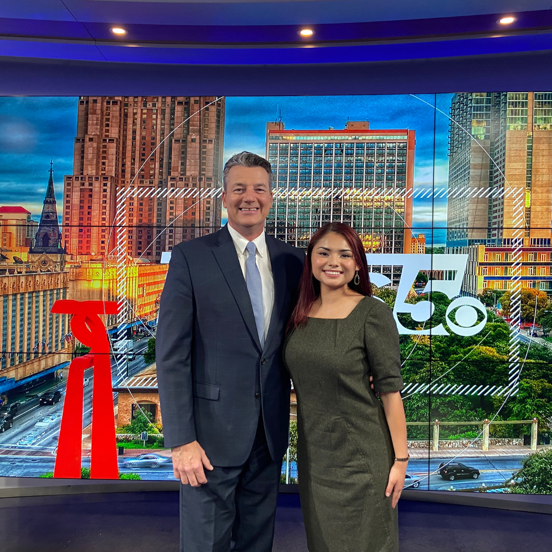 A man and a woman in business attire stand smiling together in a television news studio in front of a city skyline backdrop.