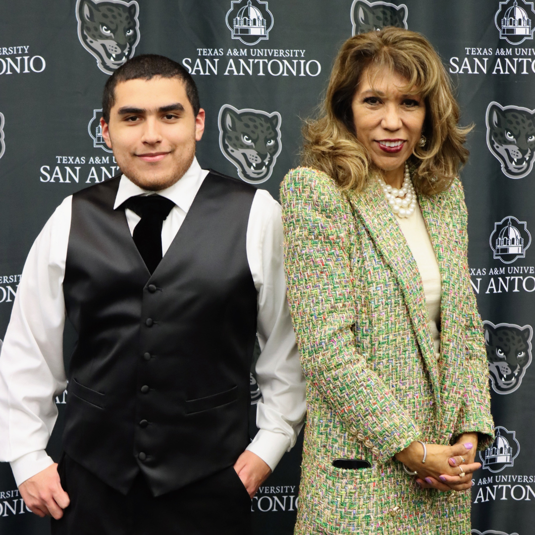 A young man in a vest and tie stands next to a woman in a green jacket before a Texas A&M University-San Antonio backdrop.